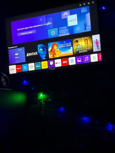 The image displays an indoor scene in Kermen, Bulgaria, dominated by a large television screen in a darkened environment. The screen is active, showing the LG webOS interface with a prominent purple banner at the top reading "Започнете едно ново изживяване с LG webOS." (Start a new experience with LG webOS.) and an "Отваряне на „Apps“" (Open "Apps") button. Below the banner, a row of streaming service application icons and content thumbnails is visible, including Netflix, Rakuten TV, Prime Video, Disney+, Twitch, YouTube, and UFC. Content promoted includes "Avatar," "Lion King II: Simba's Pride," "Dahaad," and an unspecified image featuring a person in a pink jacket on a beach. In the lower-left foreground, a rectangular object emits a strong green light horizontally. Directly beneath the television and extending into the lower-right foreground, multiple small, distinct blue light sources are scattered on an unseen surface. No individuals are present, and no specific actions or interactions are depicted.