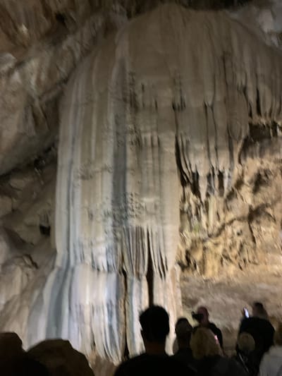 The image shows a poorly lit interior of a cave, likely in Inkiti, Abkhazia. Layered rock formations, possibly stalactites or stalagmites, dominate the scene. Several blurred figures are partially visible on the right side of the frame. They appear to be tourists or explorers. The overall environment suggests a natural cave system with geological features.