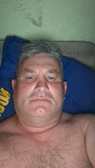 A close-up selfie of a Caucasian man with graying hair and blue eyes is shown. He is lying down on a blue pillow, with a green textured wall in the background. The man appears to be shirtless, revealing his chest hair. His expression is neutral, and he is looking directly at the camera. The image is taken from a slightly high angle, with the man's face and upper torso filling most of the frame. There is a slight sheen of sweat on his forehead and upper lip. The pillow he is lying on has a yellow design. The overall lighting suggests an indoor setting, possibly a bedroom. The location is stated to be in Voinova, Moldova.