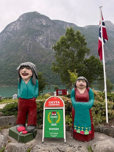 In Olden, Norway, two wooden folk art figures stand outside the Gryta Camping. The figure on the left, dressed in a teal shirt and red pants, has a large smile and grey hair. It stands on a green box with two pink stones, one inscribed with "Love." The figure on the right wears a red dress with teal sleeves and a teal apron adorned with a geometric pattern. Between them, a green and red sign displays "GRYTA CAMPING" and "ACSI AWARDS 2025 WINNER" with "Gryta Camping, Best campsite, Norway" written below. Behind the figures, a calm, turquoise body of water is visible, with a steep, tree-covered mountain range and a cloudy sky in the background. A Norwegian flag flies from a pole to the right of the figures.