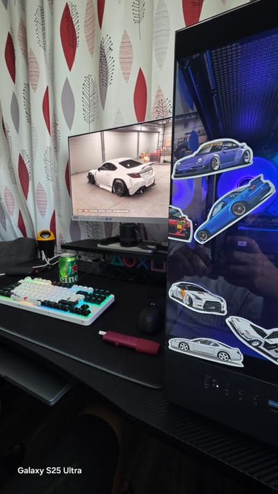 This image depicts a gaming setup, likely in a private residence. The primary focus is a desk area with a white keyboard, a black mouse, and a monitor displaying a white sports car in a garage setting. A green can of "eine" is positioned on the desk next to a black speaker. The computer tower to the right of the monitor is decorated with various car stickers, and its internal fans are illuminated with blue light. The background features a patterned curtain with red and gray leaf designs. The bottom left corner of the image displays the text "Galaxy S25 Ultra." The image does not contain information about the city of Ezerovo, Bulgaria.