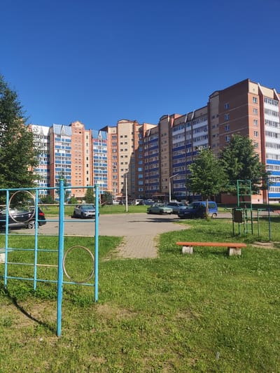 The image shows a residential courtyard in Polatsk, Belarus. In the foreground, there is a blue metal playground structure with two rings. To the right of the playground structure, a red wooden bench with concrete supports sits on a patch of green grass. Beyond the grass is a paved walkway and a parking lot with several cars. In the background, a long, multi-story apartment building with a brick facade and blue and white balconies dominates the scene. The sky is clear and blue, indicating a sunny day.