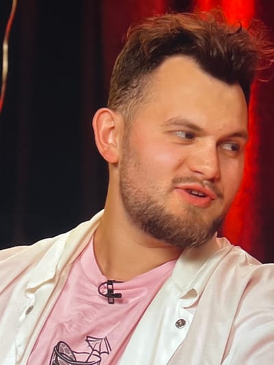 A medium shot shows a man with light brown, messy hair and a short beard. He is looking to his right, with his mouth slightly open as if speaking. He is wearing a pink t-shirt with a graphic design on the front and a white open-front jacket over it. A black microphone clip is attached to the front of his t-shirt. The background is dark, with a red curtain visible behind him. The image appears to be a still from a video recording, possibly an interview or broadcast. The location in Genemuiden, Netherlands, is not discernible from the visual information provided.