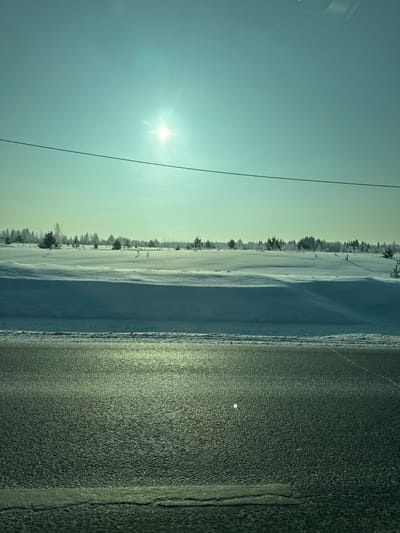 The image captures a bright, sunny winter landscape, seemingly taken from a low angle or possibly from a vehicle. The original image is oriented vertically, but the content suggests a horizontal (landscape) view.
In the upper portion of the corrected horizontal view, a clear, light blue sky dominates, extending across the frame. A brilliant, star-like sun is visible in the upper left, emitting strong rays and causing a slight lens flare effect. Closer to the horizon on the right side of the sky, the blue transitions into a faint yellowish-green hue, possibly indicating atmospheric conditions or a low sun angle.
Below the sky, a vast expanse of snow-covered ground stretches into the distance. The snow appears largely undisturbed, with subtle undulations and drifts creating texture and shadow play. Along the horizon line, a dense row of trees, primarily evergreens, stands out against the lighter sky. These trees appear to be covered with a light dusting of snow or frost, characteristic of a cold environment.
The lower and right half of the image is occupied by a wide, dark road surface. The road has a rough, granular texture, typical of asphalt, and appears somewhat wet or icy, reflecting the bright sunlight with a glossy, dark greenish-gray sheen. Several faint, irregular lines and possibly cracks are visible on its surface. A small, bright white circular object rests on the road in the middle-right area, which could be a reflective marker, a piece of debris, or an illuminated patch of ice.
A thin, dark power line or utility cable diagonally crosses the sky from the upper left corner, passing over the sun's position, and extends towards the center-right of the image.
The overall lighting is bright and crisp, characteristic of a clear winter day. The strong presence of the sun, coupled with the snow-covered environment and the icy appearance of the road, indicates cold weather conditions. There is no discernible human activity or visible text in the image.