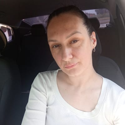 A woman is pictured inside a car, with the camera angled upwards and slightly to the left, capturing a frontal view. The woman has dark hair pulled back, and her face is lightly freckled. She is wearing a white, ribbed long-sleeved shirt. A silver hoop earring is visible on her left ear. The background is dark, with the interior of a car, including car seats, partially visible. The rear window shows a blurred view of the exterior, suggesting the car is in motion or parked. The image's metadata indicates it was taken in Petukhovo, Russia, on an unspecified date at 10:10 AM.
