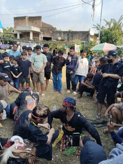 In Kedawung, Indonesia, an outdoor scene unfolds with a crowd gathered around two men in black, seated on the grass. The men are adorned with face paint and headbands. One man has blue hair. They appear to be engaged in some kind of interaction. The crowd surrounding them consists of children, teenagers, and adults, with some individuals observing and others recording the scene with their phones. In the background, buildings and trees are visible under a partly cloudy sky. A vendor's umbrella can also be seen. The grass appears dry.