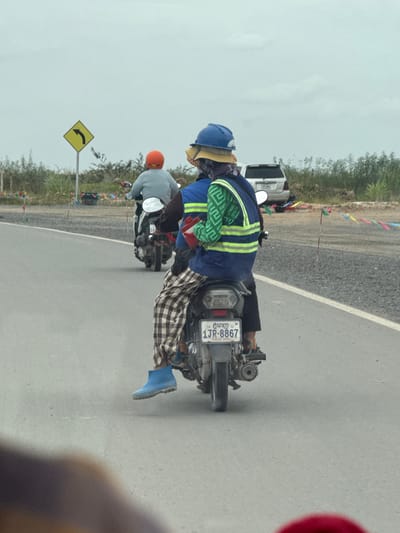 The image shows two people on a motorcycle, with the rider in front wearing an orange helmet and a light gray jacket, and the passenger behind wearing a blue helmet, a yellow hat, and a green and white patterned vest over a blue jacket. The passenger is also wearing plaid pants and blue boots. The motorcycle has a license plate that reads "1JR-8867" and "PHNOM PENH". They are on a paved road, likely in or near Krong Ta Khmau, Cambodia, given the license plate information. In the background, there is a white SUV, a yellow directional sign indicating a curve to the left, and various flags or banners strung along the side of the road. The environment appears to be a roadside with some vegetation and construction or unpaved areas visible. The sky is overcast.