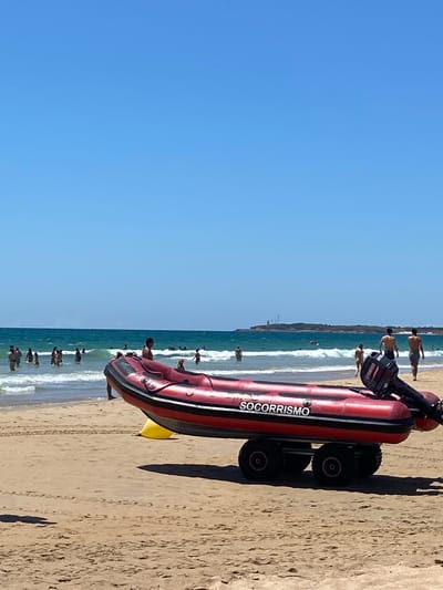 An inflatable rescue boat with the text "SOCORRISMO" is on the sandy beach in Conil de la Frontera, Spain. The boat is red with black trim and is mounted on a trailer with four wheels. A Yamaha outboard motor is attached to the stern of the boat. The sea is calm with gentle waves and a clear blue sky overhead. Several people are in the water, swimming and playing in the waves. In the background, a peninsula with some structures is visible in the distance. The scene depicts a typical summer day at the beach.