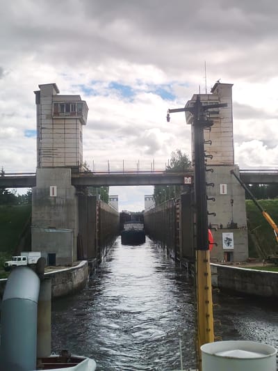 This image depicts a view of the Vytegra Lock, part of the Volga-Baltic Waterway in Vytegra, Russia. The perspective is from a boat approaching the lock gates. Two large concrete towers, characteristic of lock structures, flank the waterway, with a bridge spanning between them in the background. The lock gates themselves are closed, containing a dark-colored vessel. The water in the lock is rippled, indicating current. On the left side of the lock, a white truck is parked near a small building. The sky is overcast with some patches of blue. A prominent yellow pole with hooks extends from the foreground, suggesting it is part of the boat or the lock mechanism. The overall scene conveys industrial infrastructure and waterway navigation.