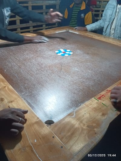 The image depicts an active carrom game in an indoor setting, identified as Maliat, Bangladesh. A wooden carrom board occupies the majority of the frame, showing a worn playing surface with a lighter, smoother center area and darker edges. The wooden frame of the board is visible, featuring a corner pocket in the lower left and faint green and red lines, along with some Bengali script, near the lower right corner.
In the center of the board, a cluster of carrom coins is arranged in a circular pattern: one red coin at the center, surrounded by one blue coin, and then encircled by multiple white coins.
Several individuals are present around the board. In the lower left foreground, a dark-skinned hand with a silver ring on the ring finger rests on the wooden frame. To the left-middle, a person's left hand, wearing a dark long-sleeved garment, is positioned over the board, appearing to be either applying powder or preparing for a strike, with the right hand slightly blurred from motion above the board. In the background, three other individuals are partially visible: one wearing a striped green shirt and a yellow and blue jacket (center-back), another wearing a light-colored jacket (right-back), and a third wearing a red cap or hood (far right-back). The background shows a slatted wooden structure, suggesting a casual, possibly open-air or semi-enclosed venue. The timestamp "03/12/2025 19:44" is visible in the bottom right corner.