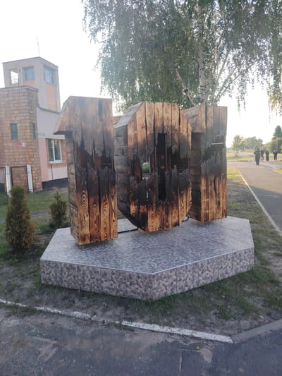 The image captures an outdoor scene in Malaryta, Belarus, featuring a large wooden sculpture resembling the number "10". The sculpture is positioned on a tiled pedestal in front of a brick building. The sculpture appears to be made from charred wooden planks, giving it a weathered and somewhat burnt appearance. Two small evergreen shrubs are planted on either side of the pedestal. To the right of the sculpture, a paved walkway is visible, and in the distance, several people are walking, one on a bicycle. The background includes trees and a clear sky, suggesting it is daytime. The building behind the sculpture has a modern architectural style with brick and light-colored sections.