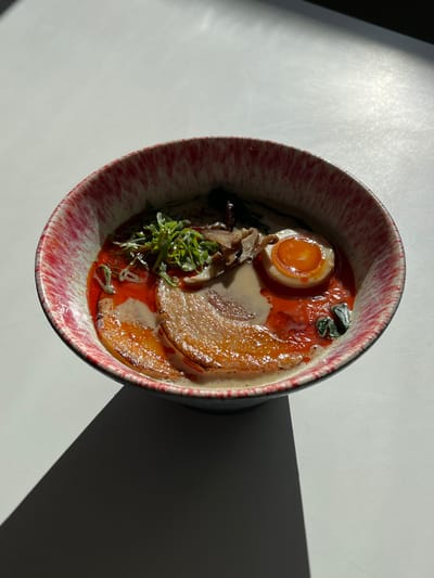 A bowl of ramen is depicted, viewed from a slight overhead angle, resting on a white, flat surface. The bowl has a dark blue base and an irregular, mottled red and white rim. Inside, the ramen features a creamy-colored broth, topped with a significant amount of bright red chili oil or paste. Visible toppings include several slices of seared pork belly (chashu), a soft-boiled egg cut in half with a vibrant orange yolk, thinly sliced green onions or chives, and what appears to be small pieces of dark mushroom or seaweed. Strong directional light from the upper right casts a dark, triangular shadow from the bowl across the lower left portion of the white surface. The scene is located in Pozuelo de Alarcón, Spain.
