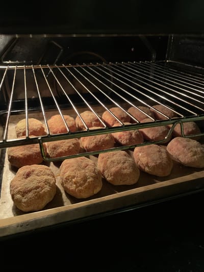 A close-up, high-angle shot shows a rack of uncooked meatballs inside an oven. The meatballs are round and covered in a light-colored breading. They are arranged in rows on a baking sheet, with some meatballs visible on the bottom rack of the oven as well. The oven interior is dark, with the shiny metal of the rack and baking sheet reflecting the light. The overall impression is one of food preparation, possibly for a meal. The image lacks any specific indicators of time of day, weather, or emotions. No visible text is present. The location context of La Línea de la Concepción, Spain, does not provide any visual cues within the image itself.