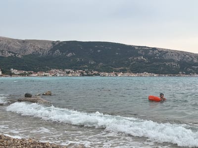 The image captures a coastal scene in Općina Baška, Croatia, under an overcast sky. In the foreground, a wave is breaking on a pebble beach, with a section of the beach visible at the bottom left. The sea stretches out into the middle ground, where a person is swimming while holding onto an orange inflatable buoy labeled "NABAIJI". The person appears to be looking towards the camera. In the background, a town with numerous buildings is nestled along the coast, backed by a large, tree-covered mountain. The mountain range extends across the horizon. The water is a muted blue-green.