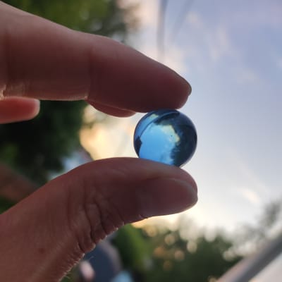 A close-up, low-angle, outdoor, eye-level shot shows a person's index finger and thumb pinching a blue glass marble against a soft-focus background of trees and sky. The sky is a pale blue with diffuse clouds and a hint of warm sunset light peeking through the trees. The marble is round and appears to be transparent, reflecting the sky and light. The fingers are pinkish-tan and detailed, showing slight wrinkling and texture. The scene is likely a park or backyard area in Kineshma, Russia, given the natural elements and soft lighting.