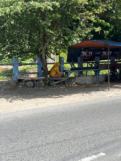 The image depicts an outdoor scene on a sunny day, likely in Ketegan, Indonesia, with a focus on a roadside area. In the foreground, a grey asphalt road with a white line on the right edge is visible. Behind the road is a dusty, sandy area with some patchy vegetation and fallen leaves. A concrete barrier runs along the edge of the road. Further back, a person wearing a yellow headscarf and brown clothing is seated on a wooden bench under a large tree. The bench is supported by large stones. Next to the bench, a broom rests against the concrete. Beyond the bench, a blue metal railing runs horizontally. Behind the railing, there is more vegetation. To the right of the tree, a stall with an orange and blue awning is set up. The stall has a dark blue banner with large white lettering that appears to read "300" and smaller text below. There are also signs on the stall displaying prices, such as "Rp 23.000". A motorcycle is parked to the right of the stall. The overall scene suggests a humble commercial or rest area by the roadside.