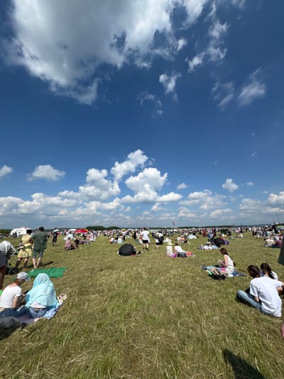 The image depicts a large outdoor gathering in Nestiukovo, Russia, on a clear, sunny day with scattered white clouds. A vast expanse of green, somewhat dry grass stretches towards the horizon, where a treeline and distant structures are visible. Numerous people are spread across the field, many seated or lying on blankets, attending an event. A variety of tents, umbrellas, and portable shelters dot the landscape, providing shade. Some individuals are standing and walking, suggesting a festive atmosphere. In the foreground, several groups are seen relaxing on the grass, including a couple sitting on a blanket, one person wearing a light blue headscarf, and another looking at a phone. Towards the middle ground, a woman in a yellow dress and a man in a green shirt are walking, and a red umbrella stands out among the crowd. The overall impression is of a busy, public event taking place in a rural or park-like setting.