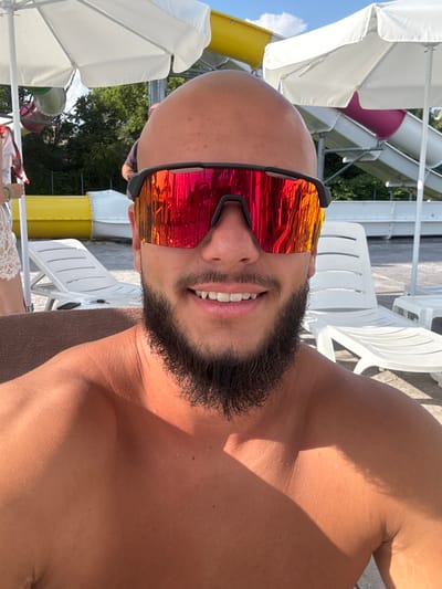 A close-up, shoulder-up selfie of a bald, bearded man wearing mirrored sunglasses with red and orange lenses. He is outdoors, possibly at a water park, with white umbrellas and water slides visible in the background. The man is shirtless, and the lighting suggests it is a sunny day. The location is identified as Ivanyane, Bulgaria.