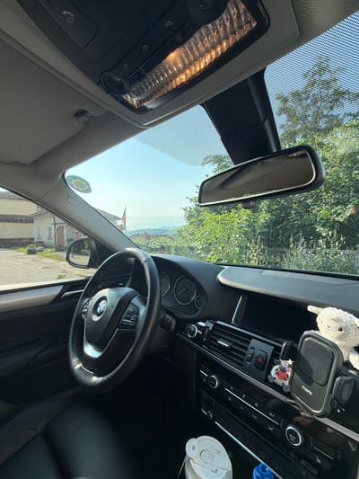The image shows the interior of a BMW car, viewed from the driver's perspective. The steering wheel, dashboard, and center console are visible. A plush toy sheep is attached to a phone holder on the dashboard. The car is parked outdoors, with a view of a rural landscape through the windshield. In the distance, there are buildings, trees, and a flag. The context suggests the location is Gorno Pavlikene, Bulgaria. The scene is well-lit, indicating it is daytime.