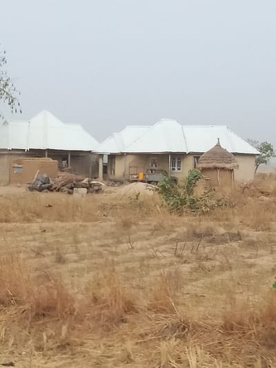 The image depicts a rural or semi-rural landscape in Minting Chip, Nigeria, under an overcast or hazy sky. The foreground consists of dry, reddish-brown earth with sparse, withered vegetation. In the midground, two primary residential structures with light-colored, corrugated metal roofs are visible. The structure on the left appears to be constructed from mud bricks, with visible construction materials including stacks of wood, black bags, and possibly mud blocks in front of it. The structure on the right is more complete, featuring plastered walls, multiple windows, and a covered veranda area where a yellow jerrycan and a dark utility vehicle or cart are discernible. To the right of this house stands a smaller, circular, mud-built hut with a conical thatched roof. No individuals are visible. The overall scene suggests a dry season environment.