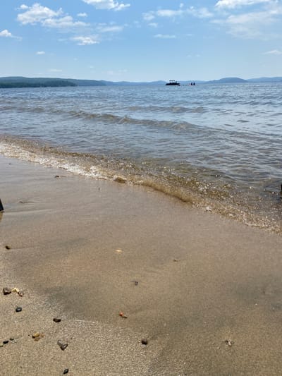 An outdoor shot captures a lake with a sandy shoreline and a distant treeline under a partly cloudy sky. The water is rippling, and a small boat with several people on board is visible in the distance, as is a pontoon boat. Several pebbles and shells are scattered across the wet sand at the edge of the water. The scene appears to be located in the municipality of Duhamel, Quebec, Canada.