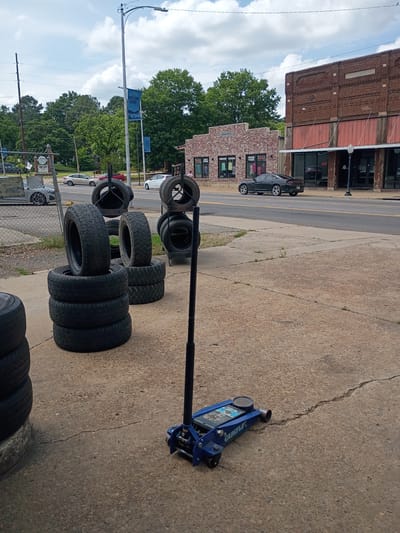 The image shows a tire shop or automotive repair establishment in Clarksville, Tennessee. Several stacks of tires of various sizes are visible on a concrete surface, along with a blue floor jack. A metal rack is holding more tires. In the background, there's a street with cars parked alongside buildings, a brick building possibly housing other retail businesses. Trees and overhead wires are visible, suggesting an urban or commercial area. The sky is partly cloudy.