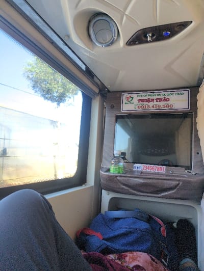 The image captures the interior of a passenger vehicle, likely a sleeper bus, viewed from a seated or reclined position. The lower-left foreground shows the bent knee and upper leg of an individual, dressed in dark gray pants. A large window on the left side of the frame provides a view of a bright outdoor environment featuring a clear blue sky, a green tree, and a light-colored building structure.
The interior ceiling above the window is light-colored and features a circular air vent and a control panel equipped with a metallic toggle switch and a blue indicator light. To the right, a dark, rectangular screen, part of a personal entertainment unit, is recessed into the pod design. Directly above this screen, a white sign with a brown frame displays Vietnamese text: "CÔNG TY CỔ PHẦN DƯỢC LIỆU THUẬN THẢO" (Thuan Thao Pharmaceutical Joint Stock Company), accompanied by a green leaf logo and the hotline number "0913.419.580".
Below the screen, a small wooden-look shelf holds a clear plastic water bottle with a green label and a white sticker marked "NK WIFI: 234567891". Beneath this shelf, an open storage compartment contains a dark blue bag with a red strap, a patterned maroon and floral blanket, and the partial view of a black shoe or foot on the far right.
The vehicle belongs to "Thuận Thảo," a company known for operating bus services that include routes to and from Dalat, Vietnam. Consequently, the scene is situated either at a bus station in Dalat or the bus is currently traversing a road within the city.