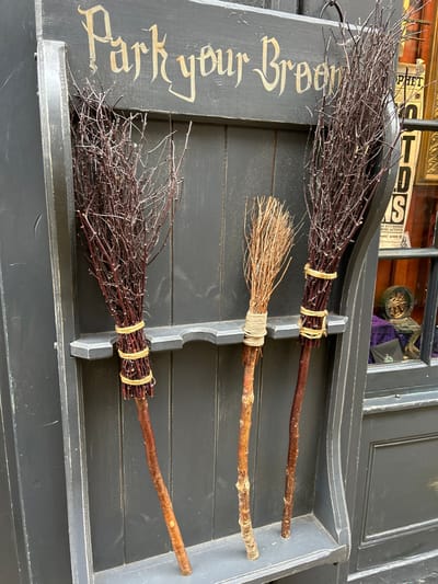Three handcrafted brooms, dark brown and straw-colored, sit in a dark gray wooden tray with the inscription "Park your Broom." The brooms feature twig bundles tied with light-colored twine to wooden handles. The tray, designed with ridges for broom placement, suggests a whimsical storage solution. The scene evokes a magical, possibly Harry Potter-themed, aesthetic.
