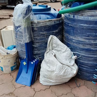 The image shows two large blue water tanks secured with plastic wrap, a pile of blue Tata shovels wrapped in plastic, a large white bag, and a white bucket. The tanks have labels indicating they are "3 Layer Heavy duty EXTRA TOUGH U.V. STABILIZED" and are ISO 9001 certified. The scene appears to be outdoors on a paved area with interlocking concrete pavers. Part of a vehicle is visible in the background. The "Fevicol MARINE" is visible on the side of the bucket. The location of the city of Oxel, India is only assumed and cannot be determined from the image.