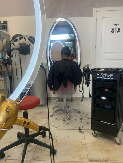 The image depicts the interior of a hair salon, likely in San Quirico d'Orcia, Italy, given the context of its surroundings. The central focus is a person seated in a salon chair, viewed from the back, with their hair being cut. They are wearing a black cape. A large, oval mirror with a bright, illuminated frame hangs on the wall behind them, reflecting the salon's interior, including another person in the background and plants.

In the foreground, a golden hair dryer is mounted on a stand, angled towards the viewer. To the right of the seated person, a black rolling cart with multiple drawers is positioned, labeled "ALFAPARF MILANO PROFESSIONAL". The cart contains various hair care tools and products. A hair dryer is also attached to the cart. The floor is tiled and scattered with clippings of hair, indicating that a haircut is in progress or has recently been completed.

On the wall to the right, there is a white door with a "WC" sign and stylized icons representing male and female figures. In the background on the left, a large, industrial-looking hair dryer with multiple vents is visible, along with shelving units and what appear to be product boxes. The overall environment suggests an active salon setting.