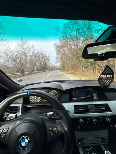 The image displays the interior perspective from the driver's seat of a BMW E60/E61 series vehicle, positioned on a rural road in Брусовeте, Bulgaria. The prominent foreground features a black leather multi-function steering wheel with a central BMW emblem, accented by a blue ring at the top of the rim, and visible silver paddle shifters. The dashboard includes an instrument cluster displaying two circular gauges and a central iDrive screen with a menu rendered in Cyrillic script. A small Baby Yoda figurine is situated on the passenger side of the dashboard. A black, leaf-shaped "areon SPORT LUX" air freshener hangs from the rearview mirror. The exterior scene, viewed through the windshield, shows a two-lane asphalt road with double yellow dividing lines, curving gently to the right. A metal guardrail parallels the left side of the road. Both sides of the road are bordered by dense clusters of deciduous trees with bare branches, indicative of a late autumn, winter, or early spring season. The sky is overcast. No individuals are visible within the vehicle or in the external environment.