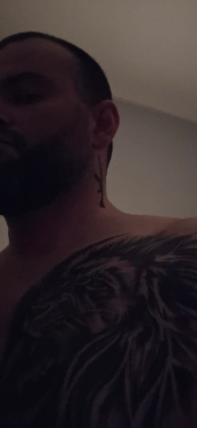 A close-up shot of a man's head and upper torso. He has dark hair and a dark tattoo of what looks like feathers on his upper left chest and shoulder. He also has a small tattoo near his ear on his neck. The background appears to be a plain, light-colored wall. No information relates to the city of Gütersloh, Germany, to be inferred from this image.