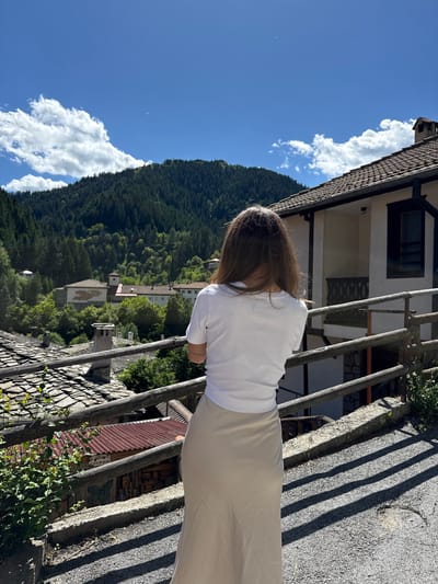 A person with long, brown hair stands with their back to the camera, looking out over a village nestled in a valley. They are wearing a white t-shirt and a long, beige skirt. In the foreground, there is a wooden railing and a stone wall. Behind the person, the village of Shiroka Laka, Bulgaria, is visible, with traditional stone houses and red-tiled roofs. The houses are surrounded by lush green trees, and a dense forest covers the mountainside in the background. The sky is a clear blue with a few scattered white clouds. The scene suggests a peaceful, rural setting on a sunny day.