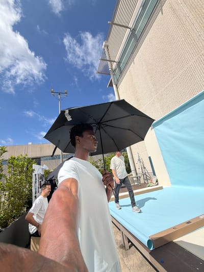 A low-angle shot captures a man holding a black umbrella. He gazes upward, toward a sky filled with scattered clouds. Adjacent to him, two people are standing near a backdrop of light blue material. The backdrop appears to be partially rolled up. The setting is outdoors, with a building in the background and trees visible on the right side of the frame. A light pole with fixtures extends into the sky. The location is Fibracolor, Spain.