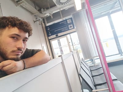 A male individual is captured in a close-up, selfie-style photograph within an indoor airport terminal, specifically identified as the German sector of EuroAirport Basel Mulhouse Freiburg, located in Saint-Louis, France. The male, with curly brown hair, a beard, and a mustache, occupies the left foreground, looking directly at the camera. He wears a dark, possibly patterned, short-sleeved shirt and a black beaded bracelet with silver accents on his left wrist. His left forearm rests on a white, modular partition.

In the mid-ground, to the right of the male, a row of airport-style seating with grey upholstery and black frames is visible. Beyond this, large, floor-to-ceiling windows offer a view of an outdoor area, indicative of an airfield. Overhead, the ceiling features exposed white ventilation ducts, electrical conduits, a circular air vent, and linear fluorescent light fixtures. A security camera is mounted near the ceiling. A blue rectangular sign, partially visible through the windows, displays German text "Gepäckausgabe" (Luggage Claim) and "Ausgang" (Exit) with downward arrows, alongside "A12-B12." A prominent, slender pink vertical structure stands on the far right of the frame. The floor is covered with dark carpeting. The scene is brightly lit by both natural light from the windows and artificial overhead lighting.