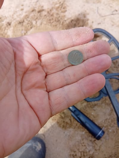 A person's hand, covered in fine sand, holds a small, tarnished coin with the number "10" clearly visible on its surface. The coin appears to be made of a greenish-bronze colored metal. In the background, a blurred sandy surface suggests an outdoor environment, possibly a beach or sandy ground. Partially visible in the background are elements of a metal detector, including a gray coil and a dark blue handle. The scene suggests the discovery of the coin using a metal detector. The approximate location is Šaukėnai, Lithuania.