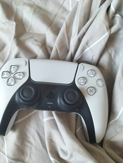 The media file shows a close-up, high-angle view of a white and black PlayStation 5 controller resting on a beige, wrinkled fabric. The fabric has thin white stripes. The controller is positioned diagonally, with its left side closer to the viewer.
The subjects are the controller itself and the fabric it lies on. The controller features a directional pad on the left, a PlayStation logo in the center, two analog sticks, and four action buttons (triangle, circle, cross, and square) on the right. There are also small buttons for power, mute, and options visible on the controller.
There is no specific activity or event taking place, as the controller is stationary. The scene is indoors, with the soft lighting and fabric suggesting a relaxed or personal setting, possibly a bed or sofa. There are no people visible in the image.
Notable details include the clean design of the controller and the texture of the fabric. The lighting appears to be natural, casting soft shadows. There are no discernible time of day or weather cues. Location context is not directly evident from the image itself, but if it is Narbonne, France, it is likely a domestic setting.
No visible text is present on the controller, aside from the standard PlayStation symbols on the buttons and the logo.