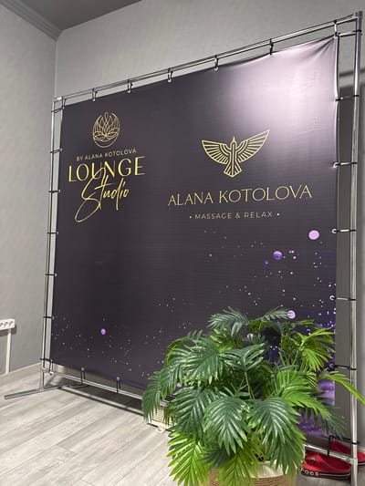 The image shows a banner with a black background and gold lettering/logos. The banner reads "BY ALANA KOTOLOVA LOUNGE Studio" in a stylized script, and "ALANA KOTOLOVA • MASSAGE & RELAX •" in a more standard font. The banner is suspended by black zip ties from a metal frame. A potted palm is in the right side of the frame, partially obscuring the banner. A pair of red clogs sit behind the banner on the right. The background shows a light grey wall on the left and a wood-paneled wall on the right. There is a white power strip is visible on the floor. The location is likely an interior setting, possibly a lounge or studio. There is no indication of Zamankul, Russia.