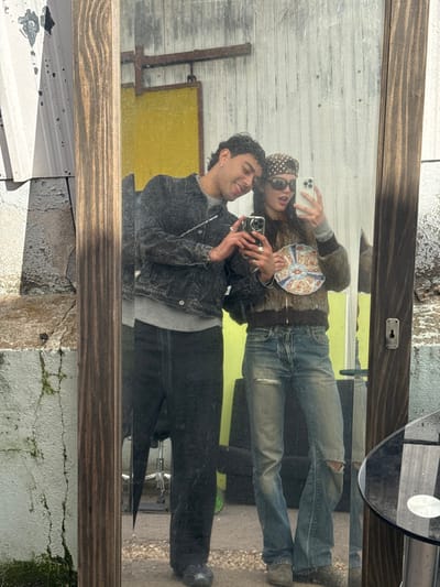 The image displays an outdoor mirror selfie featuring two young individuals in Neuilly-Plaisance, France, against a backdrop indicative of an urban industrial or salvage environment.
The male subject, positioned on the left, has dark, curly hair and wears a dark acid-wash denim jacket over a light grey crew-neck sweater, dark trousers, and dark shoes. He holds a dark-colored smartphone with both hands, positioned to photograph the reflection, and exhibits a smiling expression with his head tilted towards the female. Multiple rings are visible on his left hand, and a silver chain hangs from his jacket.
The female subject, on the right, has dark hair partially obscured by a dark, polka-dot bandana and dark sunglasses. She wears a brown textured bomber jacket over a light grey long-sleeved top, distressed blue jeans with tears at the knees, and light-colored shoes. She holds a light-colored smartphone in her left hand, also aimed at the reflection, and a circular, multicolored ceramic plate with her right hand. Her mouth is open in what appears to be a smile behind the sunglasses. Multiple rings are visible on her right hand.
The full-length, vertically oriented mirror is encased in a distressed wooden frame, featuring a keyhole plate on its right vertical side. The reflective surface exhibits minor imperfections. The background visible within the mirror's reflection includes a bright yellow wall or structure, metallic beams, and indistinct forms suggesting outdoor furniture, all resting on an asphalt or concrete surface. Outside the mirror frame, to the left, a weathered corrugated metal wall is present above a cracked concrete wall with green moss growth. To the right, a portion of a round table with a glass top and metallic base is visible in the foreground.