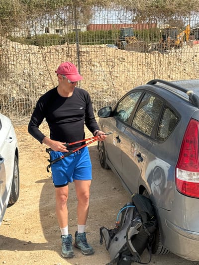 An adult male stands outdoors in the Megilot Yam HaMelakh Regional Council, Palestinian Territory, on a clear, sunny day. He is dressed in a red baseball cap with sunglasses pushed onto the brim, a black long-sleeved athletic shirt with a white 'NB' logo, blue shorts over black compression shorts, white socks, and blue/grey hiking boots. The male holds a pair of red and black collapsible trekking poles in his hands, appearing to adjust or stow them. He stands on a light brown, sandy ground between a dark grey hatchback vehicle parked to his right and a partially visible white vehicle to his left. A grey hiking backpack with a visible blue hydration tube is leaning against the right rear wheel of the dark grey car. The background consists of a tall chain-link fence topped with an overhead deterrent, behind which is a dirt embankment with sparse dry vegetation. Further in the background, a red shipping container, a white building, and two yellow heavy construction vehicles (a front-end loader and a backhoe) are visible.