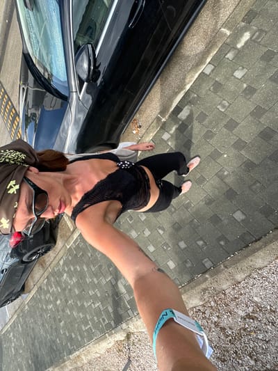 The scene, located in the city of Betina, Croatia, depicts an outdoor environment from a high-angle, slightly overhead perspective. A person stands on a paved sidewalk, facing slightly away from the camera. This individual wears dark sunglasses, a brown headscarf with light green leaf-like patterns, a black lace crop top, black trousers, and light-colored woven flat shoes. A light-colored garment is draped over their shoulders. Their right forearm is extended towards the camera, displaying a light blue and white wristband with "NIGHT" and "SELECTORS" text, and a small dark tattoo on the inner forearm.
Adjacent to the sidewalk, a black sedan is parked on an asphalt surface, reflecting the bright sky. The rear portion of a second dark vehicle is partially visible behind the sedan, showing a red taillight and a black wheel. The sidewalk consists of grey interlocking concrete pavers, some of which appear damp. A concrete curb separates the paved area from a narrow strip of gravelly ground. The scene is under strong sunlight, casting defined shadows on the pavement.