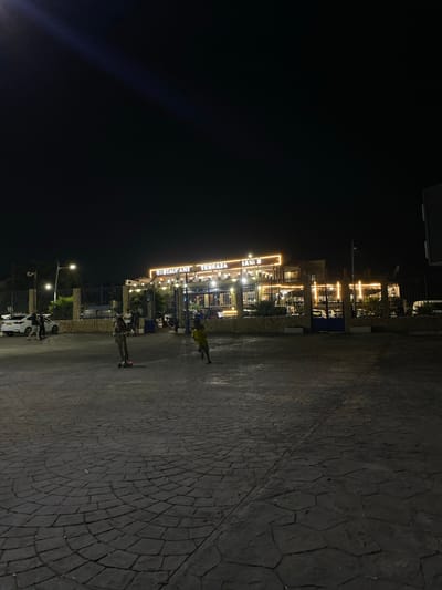 This is a nighttime outdoor photograph of a restaurant and plaza area. A building with illuminated signage reading "RESTAURANT TERRAZA JANI S" is visible in the background. The building is decorated with strings of warm-toned lights. A fence separates the plaza from the restaurant. Several vehicles, including a white car and a van, are parked in front of the building.

In the foreground, a textured paved area, possibly a plaza or courtyard, is shown. Two children are present in the plaza. One child, in the center of the frame, is riding a scooter. Another child, to the right, is running. The ground is paved with interlocking stones arranged in a circular pattern. Streetlights and building lights illuminate the scene. The sky is dark, indicating nighttime. The location is specified as Tipaza, Algeria.