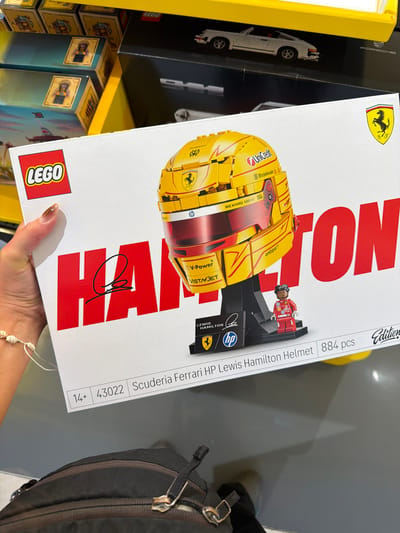 Here is a concise description of the image:
A person is holding a LEGO box for a Scuderia Ferrari HP Lewis Hamilton Helmet set in what appears to be a retail store. The box has a white background with a large LEGO logo in the lower-left corner. The helmet depicted is primarily yellow with red accents, and the text "HAMILTON" is printed in large red letters. There's also a smaller representation of Lewis Hamilton in racing gear, along with the Ferrari logo and logos of various sponsors like UniCredit, Bitdefender, and HP. The box indicates that the set contains 884 pieces and is suitable for ages 14+. In the background, there are shelves filled with other LEGO sets and a Porsche-branded box. The photo was taken in a store located in George Town, Malaysia.