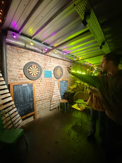 A person throws a dart towards a dartboard on a brick wall. Two dartboards and two chalkboards for scoring are visible. A xylophone leans against the wall. Green and purple string lights illuminate a metal wall. A second person is partially visible in the background. The scene appears to be in a dimly lit bar or recreation area.