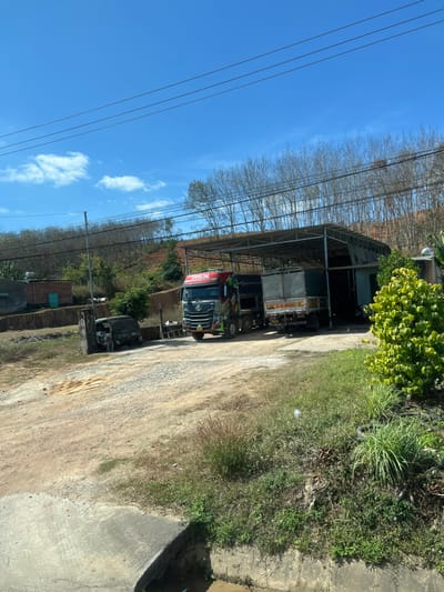 Several trucks are parked under a metal awning. Trees are on the hillside above. A dirt road leads up to the trucks. The sky is blue with sparse clouds, and power lines run across the top left. Vegetation grows along the road's edge. Concrete pavement and a structure are in the lower right. Other buildings are visible in the distance.