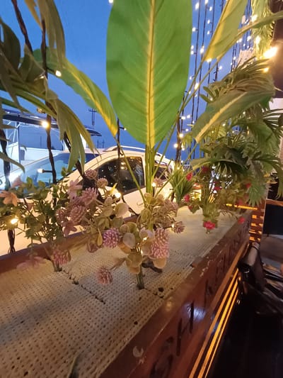 The image depicts an outdoor scene at dusk, featuring an arrangement of artificial plants in a planter box. The planter box is made of wood and contains a beige, porous material where the plants are rooted. Several types of artificial foliage are visible, including large green leaves with yellow veins, and clusters of small pink and purple flowers. String lights with warm yellow illumination are strung behind the plants, creating a festive atmosphere. In the background, a white car is parked, partially visible behind the greenery. The sky is a deep blue, indicating twilight. There is no indication of the city of Nematjan, Uzbekistan in the image.