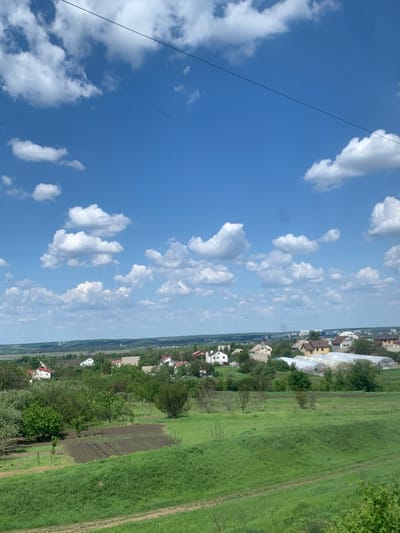 The image shows a panoramic view of a suburban area in Korotych, Ukraine, located at latitude 49.956 and longitude 36.031. The scene is dominated by a bright blue sky with scattered white cumulus clouds. In the foreground, a rolling green field with a dirt path stretches towards a cluster of trees and houses. The houses are mostly single-story structures with visible white walls and red roofs. A few greenhouses can be seen in the distance, suggesting agricultural activity. There are no people present in the scene. The overall atmosphere is peaceful and idyllic.