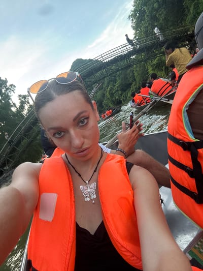 A woman is taking a selfie in a boat on the water, wearing a bright orange life vest and sunglasses. She has on a butterfly pendant necklace. In the background, other people in life vests are also in boats, and a wooden structure resembling a bridge or walkway is visible over a lush green, treed hillside. The scene appears to be a tourist activity on a river or lake in a natural setting. The presence of boats, life vests, and the mountainous terrain suggests this location could be near the karst landscape of Ninh Bình province, possibly within Yên Sơn Ward, Vietnam.
