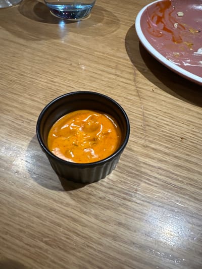 A small, black ceramic fluted ramekin bowl, approximately 7-8 cm in diameter, containing an orange-red creamy sauce, is centrally positioned on a light brown wooden table surface. The sauce exhibits a textured appearance with darker red or brown flecks, consistent with herbs or spices. In the upper left quadrant, a partial view of a clear glass filled with a translucent light blue liquid is visible. To the upper right, a segment of a round ceramic plate with a distinct pink rim and a white underside is present. This plate displays remnants of reddish sauce and small food particles, indicating recent use. Shadows are cast to the lower right of the ramekin and below the glass, suggesting an overhead light source. The scene depicts a dining context, likely within a restaurant or eatery located in Kalyan-Dombivli, India. The precise address or specific establishment within Kalyan-Dombivli cannot be determined from the visual evidence provided.