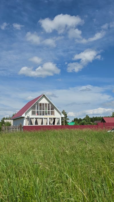 The image depicts a house with a red roof and white siding in a rural setting. The house has a large gable window on the upper floor and several windows on the lower floor, some of which are covered by curtains. A red fence runs in front of the house, separating it from a field of tall green grass. In the background, there are trees and other houses, suggesting a village or small settlement. The sky is blue with scattered white clouds. The scene is peaceful and appears to be a sunny day. The location is identified as Kupoyarovo, Russia.